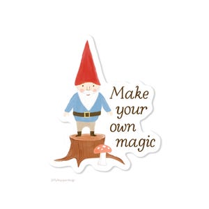 May include: A die-cut sticker featuring a gnome with a red hat, blue shirt, and brown belt standing on a brown tree stump next to a small red and white mushroom. The text "Make your own magic" is printed in brown script to the right of the gnome.