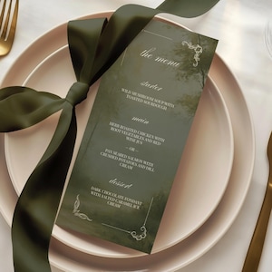 May include: A rectangular menu card with a dark green forest-themed background and white text, tied with a dark green satin ribbon. The menu is placed on a light pink plate, accompanied by a gold fork and a gold knife on a white surface.