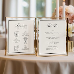 May include: Two vertical rectangular signs in thin gold metal frames. The left sign is titled Signature Cocktails with illustrations of an Old Fashioned, Espresso Martini, and Negroni. The right sign is titled The Bar, listing wine, spirits, beer, and non-alcoholic options in black text on a cream background.