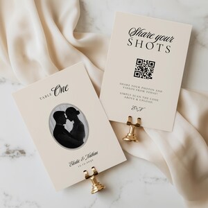 May include: Two cream-colored rectangular cards held by gold metal stands. One card reads "TABLE One" with an oval black and white photo of a couple and the text "Elodie & Nathan 12.12.2030". The other card reads "Share your SHOTS" with a QR code.