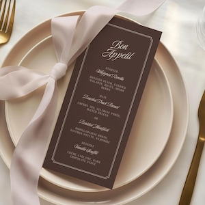 May include: A dark brown rectangular menu card with white text reading Bon Appetit, Starter Whipped Ricotta, Main Roasted Duck Breast or Roasted Monkfish, and Dessert Vanilla Souffle, tied with a pale pink ribbon on a cream-colored plate.
