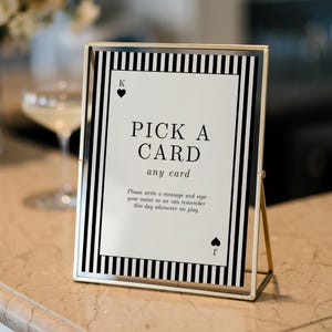 May include: A gold-toned metal frame holding a white sign with black vertical stripes. The sign features a playing card design with the text: PICK A CARD any card. Please write a message and sign your name so we can remember this day whenever we play.