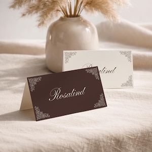 Puede incluir: Un tarjetón de mesa plegado de color marrón oscuro con el nombre Rosalind en cursiva blanca y detalles de filigrana blanca en las esquinas, situado frente a otro tarjetón crema con adornos y texto a juego, ambos con una tipografía elegante.