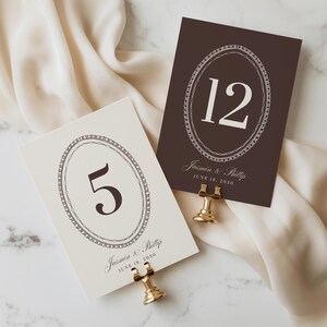 Puede incluir: Dos tarjetas rectangulares de número de mesa sostenidas por soportes de metal dorado. Una tarjeta es crema con el número 5, la otra es marrón oscuro con el número 12. Ambas presentan un diseño de marco ovalado y el texto Jasmin & Phillip JUNE 18, 2030.