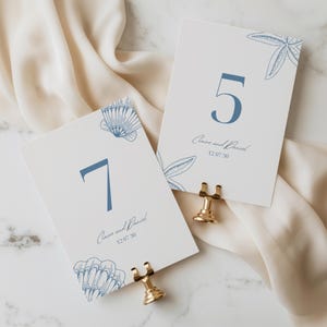 May include: Two white rectangular table number cards featuring blue numbers 5 and 7, blue line-art illustrations of seashells and a starfish, and the printed text Claire and Daniel 12 07 30. Each card is held upright by a gold metal stand.