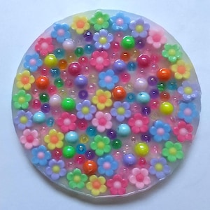 May include: A round, colorful resin coaster with embedded flowers and beads. The coaster features a variety of small, plastic flowers in shades of pink, blue, yellow, and green, interspersed with colorful beads. The overall design is bright and cheerful.