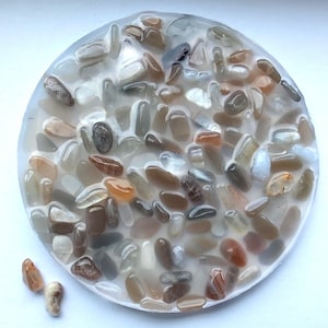 May include: A round, clear resin coaster filled with a variety of small, polished stones. The stones are in shades of brown, gray, and orange, creating a natural, earthy aesthetic. A few loose stones are scattered around the coaster.