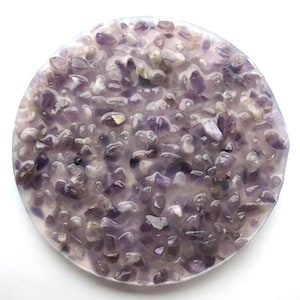 Op de afbeelding: Een ronde onderzetter met een oppervlak van kleine, paarse amethiststenen in heldere hars. De onderzetter heeft een lichtgekleurde, licht verhoogde rand. De amethiststenen variëren in grootte en tint.