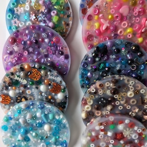 Puede incluir: Un conjunto de posavasos redondos de resina, incrustados con pequeñas cuentas, lentejuelas y elementos decorativos. Los colores varían, incluyendo tonos de azul, rosa y morado. Los posavasos están dispuestos verticalmente.