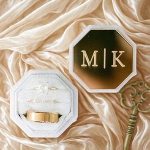 May include: Wedding ring set in a white velvet octagonal box, with a gold band and two diamond rings. A second gold octagonal box displays the initials "M|K". The background is a draped, cream-colored fabric, with a gold key.