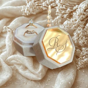 May include: A grey velvet ring box with a gold interior and engraved initials, open to reveal two rings. One ring features a pear-shaped stone, the other has a row of small stones. The box sits on a white, textured fabric.