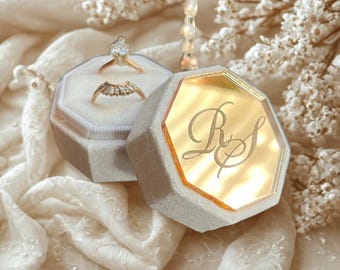 May include: A gray velvet ring box with a gold interior and engraved initials, open to reveal two rings. One ring features a pear-shaped stone, the other has a row of small stones. The box sits on a white, textured fabric.