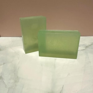 Pode incluir: Duas barras retangulares de sabão verde translúcido. Uma barra está em pé e a outra está deitada horizontalmente. As barras de sabão estão sobre uma superfície marmorizada branca e cinza, com um fundo rosa.