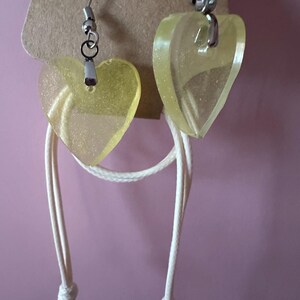 May include: A pair of heart-shaped earrings with a pale yellow, translucent appearance. The earrings are suspended from silver-colored hooks and attached to a white cord. The earrings are displayed against a pink background.