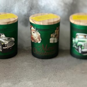 May include: Three green glass candle jars with wooden lids. Each jar features a different vintage Christmas-themed graphic: a classic car, Santa Claus, and a pickup truck. The middle jar has "Merry Christmas" text.