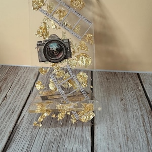 Puede incluir: Un bloque de resina transparente con una imagen de cámara vintage, detalles de tira de película y detalles de pan de oro. La cámara tiene una lente detallada. El pan de oro añade un toque de elegancia a la obra de arte.