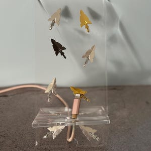 May include: A display of decorative arrow-shaped USB charging cables in silver, gold, and black. The cables are arranged on a clear acrylic stand, showcasing the unique arrowhead designs. The background is a neutral gray.