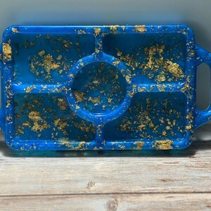 May include: A rectangular blue resin tray with a divided design and handles. The tray is adorned with gold leaf flakes, creating a decorative and functional piece. The tray has a central circular compartment and four smaller rectangular compartments.
