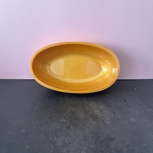 Könnte beinhalten: Eine kleine, ovale gelbe Keramikschale. Die Schale hat einen glatten, glänzenden Innenraum und eine leicht strukturierte Außenseite. Die Schale ist leer und steht auf einer dunklen Oberfläche mit hellviolettem Hintergrund.