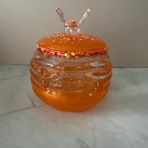 Könnte beinhalten: Ein rundes, orangefarbenes, durchscheinendes Harzgefäß mit einer an einen Bienenstock erinnernden Rippenstruktur. Der Deckel hat einen klaren, geformten Bienengriff und ist am Rand sowie auf der Oberseite mit orangefarbenem und rosa Glitzer verziert.