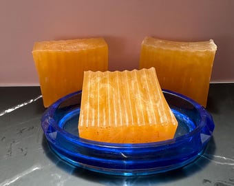 May include: Three bars of orange soap, one in a blue glass soap dish. The soap has a textured, ridged surface. The soap dish is round with a decorative edge. The background is a solid pink colour.