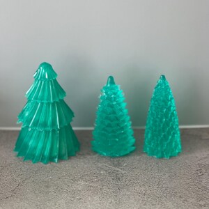 May include: Three translucent teal Christmas tree decorations. The trees vary in size and design, with one having tiered layers, another a more conical shape, and the third a simple cone. They are arranged on a grey surface.