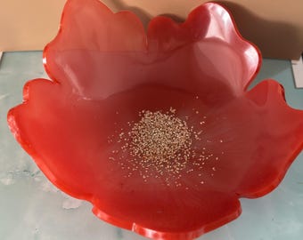 May include: A vibrant red, flower-shaped resin bowl. The bowl has a glossy finish and contains a scattering of small, light-colored granules in the center. The edges of the bowl are gently scalloped.