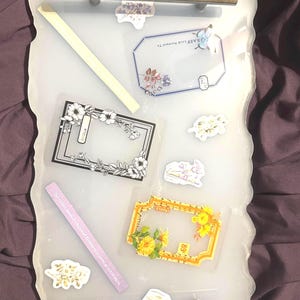 Puede incluir: Una bandeja decorativa con asas doradas y un borde blanco irregular. La bandeja contiene varias pegatinas y etiquetas con diseños florales y texto. Las pegatinas incluyen una etiqueta rectangular con el texto "GRASP Look Forward To".
