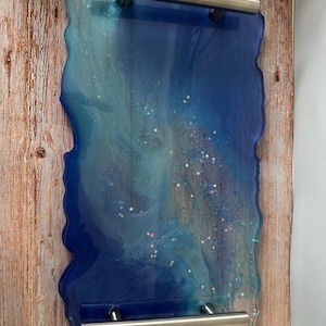 Puede incluir: Bandeja rectangular de resina con borde ondulado, con tonos azules y purpurina plateada. La bandeja tiene dos asas plateadas y está sobre un fondo de madera. La bandeja mide aproximadamente 30 cm de largo y 20 cm de ancho.