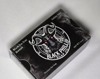 Pode incluir: Uma caixa preta de sabonete em barra premium "Black Phillip Soap Co.". A caixa apresenta um gráfico de uma cabeça de cabra com olhos vermelhos dentro de um design circular. A caixa é rotulada como "Black Tea Tree Scented" e "NET WT. 04 oz."