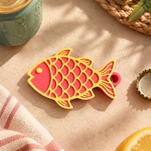 May include: A flat, fish-shaped bottle opener made of red silicone with a bright yellow outline and scale pattern. The design features a circular hole at the tail end for hanging and a small yellow dot representing the eye on the fish head.