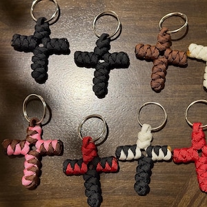 May include: A collection of handmade cross keychains in various colors, including black, brown, white, pink, red, and a black and white combination. Each cross is crafted from woven cord and attached to a silver keyring.