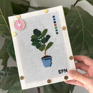 May include: A white needlepoint canvas featuring a cross-stitched fiddle leaf fig plant in a blue pot, a color palette strip, and the letters BPN. A pink donut-shaped needle minder is attached to the top left, secured by gold-toned metal magnets.