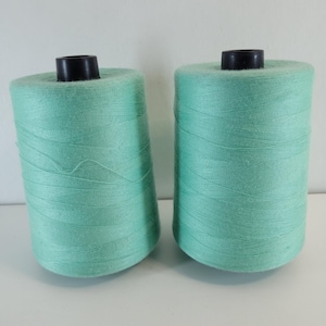May include: Two large cylindrical spools of mint green sewing thread. Each spool features a solid black plastic core at the top and bottom, with the mint green thread wound tightly around the central column in a smooth, consistent pattern.