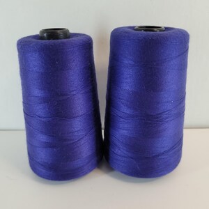 May include: Two cylindrical spools of vibrant purple thread, each featuring a smooth, tightly wound surface and a black plastic core at the top and bottom. The thread has a slight sheen and is wrapped evenly around the central spool structure.