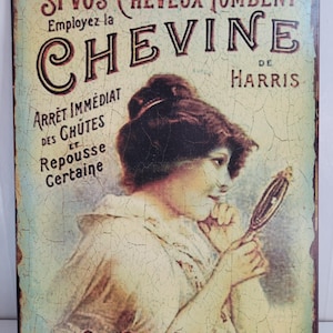 Puede incluir: Letrero metálico de estilo vintage con texto en francés. Presenta una ilustración de una mujer mirándose en un espejo de mano. El texto anuncia "CHEVINE" para la caída del cabello, con "EN VENTE ICI" en la parte inferior.