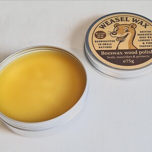 K&ouml;nnte beinhalten: Nahaufnahme einer Dose Weasel Wax Bienenwachs-Holzpolitur. Die Dose enth&auml;lt ein glattes, gelbes Wachs. Der Deckel, daneben platziert, zeigt einen Cartoon-Wiesel und den Text "Beeswax wood polish" und "e75g".