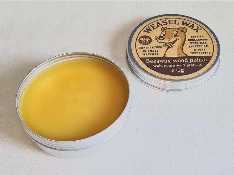 K&ouml;nnte beinhalten: Nahaufnahme einer Dose Weasel Wax Bienenwachs-Holzpolitur. Die Dose enth&auml;lt ein glattes, gelbes Wachs. Der Deckel, daneben platziert, zeigt einen Cartoon-Wiesel und den Text "Beeswax wood polish" und "e75g".