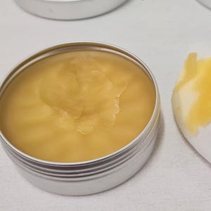 K&ouml;nnte beinhalten: Eine kleine, runde, silberne Dose gef&uuml;llt mit einem glatten, gelben Balsam. Ein wei&szlig;es Tuch mit einem Klecks des Balsams befindet sich neben der Dose. Der Balsam hat eine leicht strukturierte Oberfl&auml;che. Der Hintergrund ist eine wei&szlig;e Oberfl&auml;che.