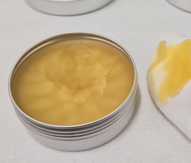 K&ouml;nnte beinhalten: Eine kleine, runde, silberne Dose gef&uuml;llt mit einem glatten, gelben Balsam. Ein wei&szlig;es Tuch mit einem Klecks des Balsams befindet sich neben der Dose. Der Balsam hat eine leicht strukturierte Oberfl&auml;che. Der Hintergrund ist eine wei&szlig;e Oberfl&auml;che.