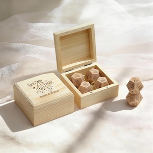 Peut inclure: Deux boîtes en bois, une ouverte et une fermée, avec des détails gravés. Le couvercle de la boîte fermée indique "Date Night" avec des cerises et des dés, et les noms "Emily & Robert". La boîte ouverte contient des dés en bois. Une pile de dés est également visible.