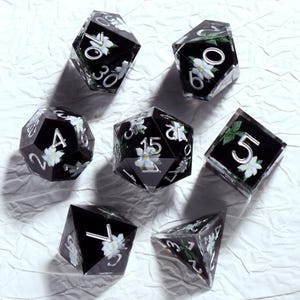 Puede incluir: Conjunto de siete dados poliédricos con fondo negro y diseños florales blancos. Los dados, con numeración plateada, tienen varias formas geométricas, incluyendo un d4, d6, d8, d10, d12, d20 y un dado de porcentaje.