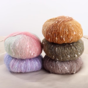 May include: A stack of five colorful yarn balls. The yarn balls are in shades of pink, blue, green, brown, and purple. The yarn has a soft, fluffy texture with white flecks throughout. The yarn is stacked on a light beige surface.