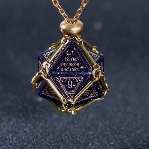 May include: A gold-colored necklace featuring a dark blue, multi-sided pendant with gold accents. The pendant has the inscription "You're my moon and stars." and the number 8. The necklace is suspended from a delicate gold chain.