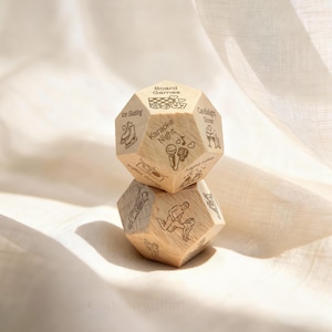 Peut inclure: Deux dés en bois à multiples faces empilés. Chaque face présente une activité différente, comme "Jeux de société", "Soirée karaoké" et "Patinoire". Les dés sont de couleur brun clair avec des illustrations noires gravées.