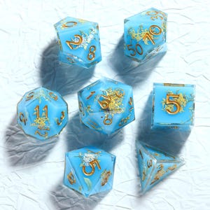 Puede incluir: Un conjunto de siete dados poliédricos de varias formas. Los dados son de color azul claro translúcido con números y diseños florales dorados. Los dados están dispuestos sobre una superficie blanca.