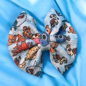 May include: A light blue fabric bow featuring cartoon characters from the movie "Finding Nemo". The bow has a variety of sea creatures printed on it, including sharks, turtles, and clownfish. The bow is tied in the center.