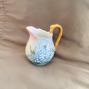 May include: A small, hand-painted porcelain creamer with a gold-colored handle. The creamer is decorated with blue and white hydrangea flowers and has a pink and white rim. The handle has a bamboo-like design.
