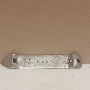 May include: A silver bottle opener with the word "HANDY" engraved on it. The opener has a rectangular shape with a pointed end and a hole on the opposite end. The word "HANDY" is centered on the opener.