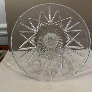 May include: A clear glass cake stand featuring a circular top with a prominent starburst pattern radiating from the center. The design includes deep, sharp-edged cuts that form a large star shape, complemented by a smaller, intricate sunburst motif.